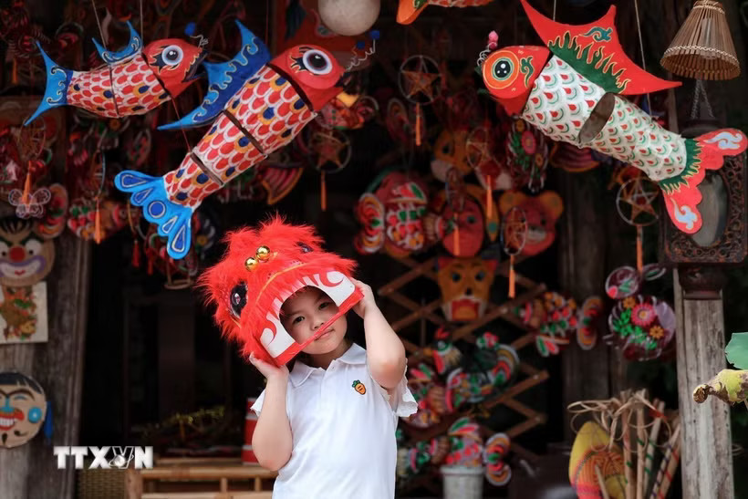Em nhỏ thích thú đội đầu lân giữa không gian rực rỡ của đèn cá chép, mặt nạ và đồ chơi Trung thu truyền thống. (Ảnh: Phạm Tuấn Anh/TTXVN)